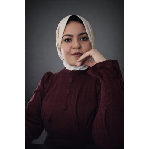 Portrait of a woman wearing a white hijab and maroon cardigan, resting her chin on her hand against a dark gray background